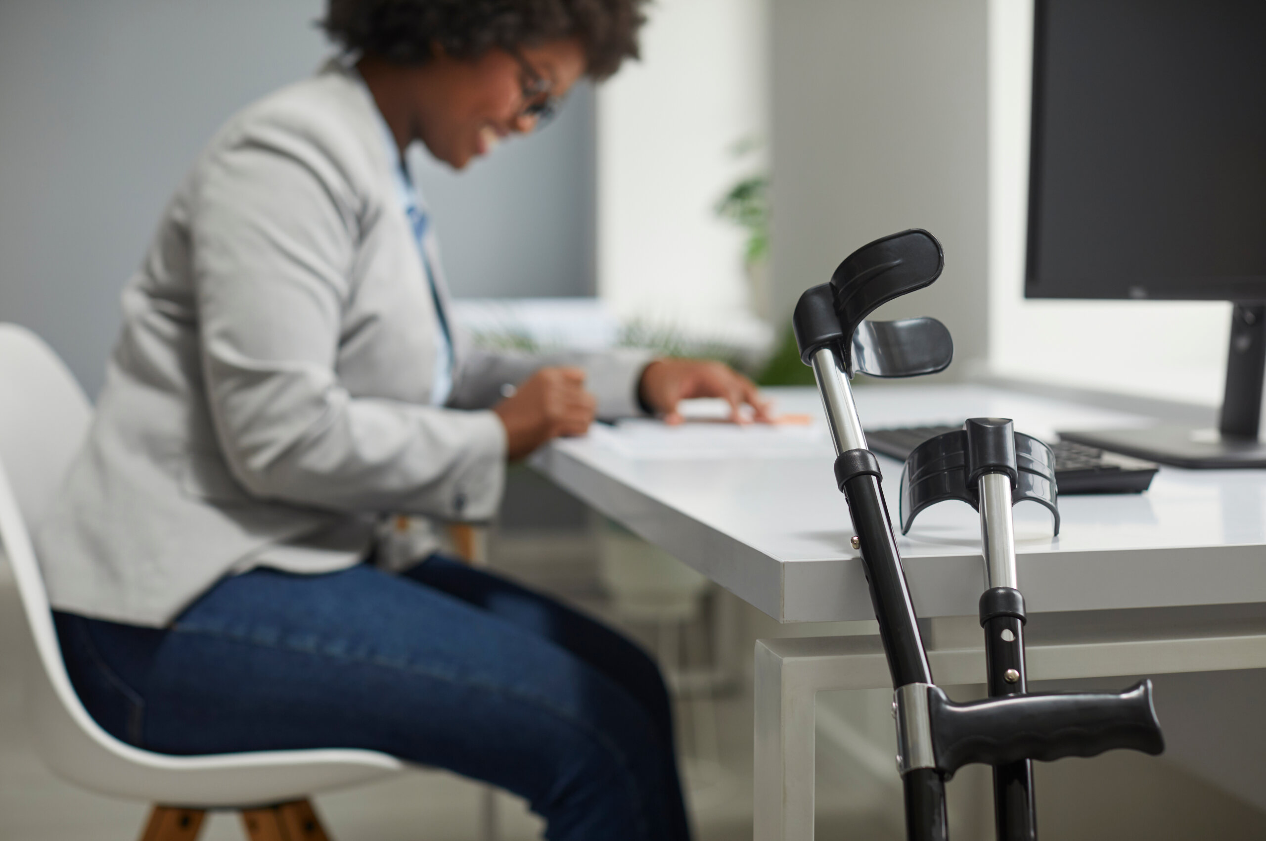 Disabled employee working in office. Orthopedic elbow crutches leaning on desk, with happy disabled African American woman working on computer in background. Working with disability concept